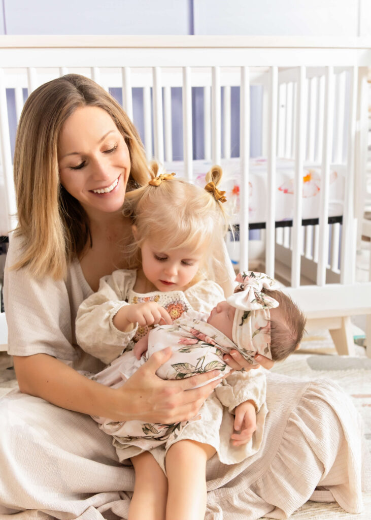 Mother sitting in a nursery with her toddler and newborn, all wearing outfits in cream and sage green floral patterns. The room features a lilac wall, creating a cohesive look with the Lilac & Earth color palette.