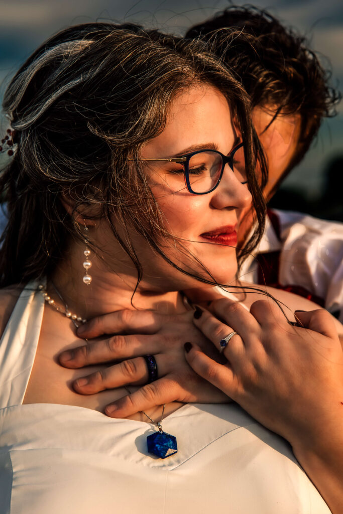 Bride with glasses and pearl jewelry being embraced by her partner at golden hour, showing wedding rings and a blue crystal necklace. A romantic and emotionally rich wedding portrait highlighting authentic connection.